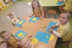 Przy stoliku grupa dzieci . Dzieci wyklejają słoneczka z koła i żółtych pasków papieru na niebieskiej kartce kartonu.