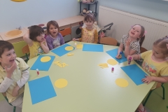 Przy stoliku grupa dzieci . Dzieci wyklejają słoneczka z koła i żółtych pasków papieru na niebieskiej kartce kartonu.