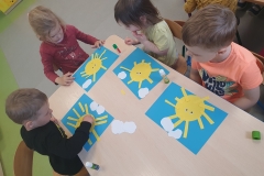 Przy stoliku grupa dzieci . Dzieci wyklejają słoneczka z koła i żółtych pasków papieru na niebieskiej kartce kartonu.