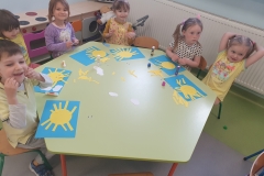 Przy stoliku grupa dzieci . Dzieci wyklejają słoneczka z koła i żółtych pasków papieru na niebieskiej kartce kartonu.