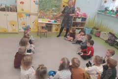 Pani w fioletowo- popielatej sukience i czarnych rajstopach stoi przy obrazkowej mapie Polski. Żółtym długopisem wskazuje na mapie napis: Kraków. Obok mapy stoi krzesełko z fioletowymi nogami. W półkolu na szarej podłodze siedzą dzieci, skierowane twarzami w stronę mapy.