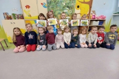 Chłopczyk w brązowym dresie i dziewczynka w białej bluzce wychylają się ze smoczej jamy zrobionej z kartonu i szarego papieru.Obok nich na podłodze siedzi 7 dzieci. Za jamą ustawiony mur z żółtych, niebieskich, zielonych i czerwonych klocków Za nim stoi 7 dzieci.Koło ściany beżowa meblościanka. W tle okno, na parapecie kolorowe kwiaty.