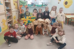 11 dzieci siedzi na szarej podłodze. Za nimi stoi 5 dzieci i trzyma w rękach obrazki historyjki o smoku wawelskim. W tle beżowe szafki z kolorowymi malowidłami oraz obrazkowa mapa Polski.