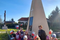Grupa dzieci z wychowawczynią stoi przed pomnikiem żołnierzy poległych w czasie ll wojny światowej. W rękach trzymają flagi Polski.