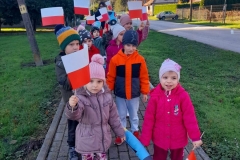Grupa dzieci idzie chodnikiem trzymając się przedszkolnego węża, w rękach trzymają flagi Polski.