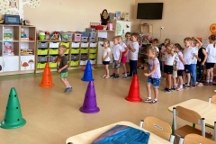 Dzieci stoją w dwóch rzędach, dziewczynka I chłopczyk biegną między kolorowymi pachołkami.