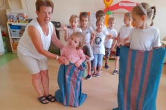 Dwie dziewczynki skaczą w workach, jednej pomaga wychowawczyni, w tle dwa rzędy dzieci.