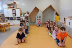 Sala przedszkolna . Grupa dzieci ubrana na kolorowa siedzi na podłodze w dwóch szeregach .Dzieci uśmiechnięte patrzą na wprost. Za dziećmi w tle mebelki przedszkolne z zabawkami i układankami .Z lewej strony widać część stolika