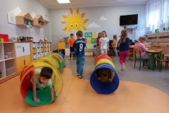 Dzieci ustawione w dwóch szeregach jedno za drugim przechodzą przez kolorowy tunel. Dwoje dzieci zwrócone tyłem po przejściu przez tunel wraca na koniec szeregu.W tle za dziećmi biała tablica z napisem DZIEŃ SPORTU , na ścianie wymalowane duże słoneczko i 2 chmurki. Z prawej strony widać telewizor , część stolika dla dzieci i biurko nauczyciela , z lewej strony szafki indywidualne dla dzieci, oraz wiszące trójkątne półki z pluszakami. Z prawej strony przy stoliku siedzi dziewczynka w rózowej bluzeczce i układa drewniane puzzle.