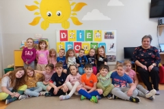 Dzieci ustawione w dwóch rzędach . Dzieci z pierwszego rzędu siedzą w półkolu na podłodze , a dzieci z drugiego rzędu siedzą na ławeczce . Po prawej stronie na krzesełku siedzi dorosła osoba- wychowawczyni . Dzieci się uśmiechają. Za dziecmi w tle biała tablica z literkami naklejonymi na kolorowe prostąkatne kartki tworzą napis DZIEŃ SPORTU . Nad tablicą na scianie widac duże namalowane słoneczko.