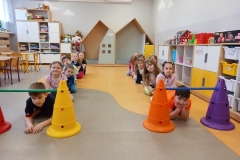 Sala przedszkolna . Grupa dzieci ubrana na kolorowa siedzi na podłodze w dwóch szeregach . Dwoje chłopców przechodzi pod przeszkodą – mostkiem z dwóch pachołków i laski gimnastycznej. W tle mebelki przedszkolne . Z lewej strony widać dwa stoliki , z prawej szafki na półki indywidualne . Na pólkach leżą poukładane klocki i zbudowana z klocków straż