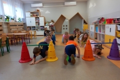 Sala przedszkolna . Grupa dzieci ubrana na kolorowa siedzi na podłodze w dwóch szeregach . Dwoje dzieci ; chłopiec i dziewczynka przechodzą po mostkiem. Jeden chłopiec odwrócony tyłem w klęku. Między szeregami na środku klęczy dziewczynka ubrana w różową bluzeczkę z twarzą zwróconą w kierunku chłopca , który klęczy tułem. Z lewej strony widać trzy stoliki , z prawej szafki na półki indywidualne . Na pólkach leżą poukładane klocki i zbudowana z klocków straż