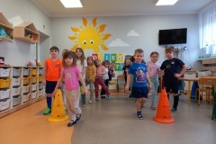 Sala przedszkolna . Dzieci ubrane na kolorowo stoją w dwóch szeregach . Przed każdym szeregiem pachołek Dwoje dzieci; chłopczyk i dziewczynka którzy stoją pierwsi w szeregu skaczą na jednej nodze . W tle za dziećmi biała tablica z napisem DZIEŃ SPORTU , na ścianie wymalowane duże słoneczko i 2 chmurki. Z prawej strony widać telewizor , część stolika dla dzieci i biurko nauczyciela , z lewej strony szafki indywidualne dla dzieci, oraz wiszące trójkątne półki z pluszakami.