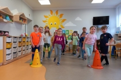 Sala przedszkolna . Dzieci ubrane na kolorowo stoją dwóch szeregach . Przed każdym szeregiem pachołek Dwie dziewczynki które stoją pierwsze skaczą na jednej nodze . W tle za dziećmi biała tablica z napisem DZIEŃ SPORTU , na ścianie wymalowane duże słoneczko i 2 chmurki. Z prawej strony widać telewizor , część stolika dla dzieci i biurko nauczyciela , z lewej strony szafki indywidualne dla dzieci, oraz wiszące trójkątne półki z pluszakami.