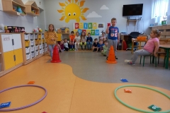 Dzieci ustawione w dwóch szeregach , kilkoro dzieci siedzi na ławeczce pod białą tablicą z napisem DZIEŃ SPORTU .Przed każdym szeregiem pachołek . Dwoje dzieci ; chłopiec i dziewczynka rzucają woreczkami do obręczy. Z prawej strony dziewczynka w różowej bluzeczce siedzi na krzesełku.