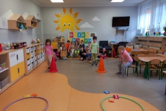 Dzieci ustawione w dwóch szeregach , kilkoro dzieci siedzi na ławeczce pod białą tablicą z napisem DZIEŃ SPORTU .Przed każdym szeregiem pachołek . Dwoje dzieci ; chłopiec z prawej strony i dziewczynka z dziewczynka z lewej rzucają woreczkami do obręczy. Z prawej strony dziewczynka w różowej bluzeczce siedzi na krzesełku.