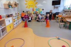 Dzieci ustawione w dwóch szeregach , kilkoro dzieci siedzi na ławeczce pod białą tablicą z napisem DZIEŃ SPORTU .Przed każdym szeregiem pachołek . Dwie dziewczynki rzucają woreczkami do obręczy. Z prawej strony dziewczynka z dwoma kucykami i w czarnej bluzeczce z napisem NIKE stoi przy biurku nauczyciela.