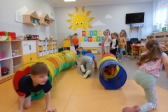 Dzieci ustawione w dwóch szeregach jedno za drugim przechodzą przez kolorowy tunel.W tle za dziećmi biała tablica z napisem DZIEŃ SPORTU , na ścianie wymalowane duże słoneczko i 2 chmurki. Z prawej strony widać telewizor , część stolika dla dzieci i biurko nauczyciela , z lewej strony szafki indywidualne dla dzieci, oraz wiszące trójkątne półki z pluszakami.