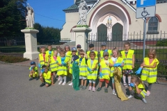 Grupa dzieci ubrana w kamizelki odblaskowe stoi w dwóch rzędach.  Przedszkolaki mają ubrane niebieskie rękawiczki, trzymają kolorowe worki do segregowania śmieci. W tle jest ogrodzenie metalowe oraz dwie figury przedstawiające postacie świętych.