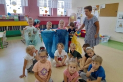 Na zdjęciu grupa dzieci siedzi na podłodze dwóch chłopców i dziewczynka trzymają w ręku kolorowe worki na śmieci. W tle wychowawczyni tłumaczy co wrzucamy do poszczególnych worków.