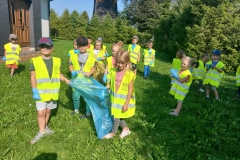 Grupa dziewczynek i chłopcow trzyma niebeiski worek. Dzieci rozglądają się w poszukiwaniu porzuconych śmieci na trawniku