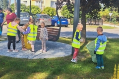 Dzieci ubrane w kamizelki odblaskowe zbierają śmieci  na trawniku. W tle ławka i samochody.