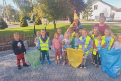 Grupa dzieci stoi  w półkolu  na szarej kostce. Za nimi stoi pani w bordowej bluzce oraz eko- skrzat w żółtej czapce na głowie.  Dzieci ubrane w kamizelki odblaskowe trzymają worki na śmieci: zielony, żółty i niebieski.