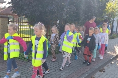 Dzieci ubrane w kamizelki odblaskowe idą w parach po chodniku. Za nimi  idzie pani. W tle zielone drzewo i metalowy płot.