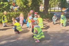 Chłopiec w odblaskiwej kamizelce odwrócony plecami trzyma zielony worek. Przy nim kolega w szarym dresie pochyla się. Obok nich grupa dzieci zbiera śmieci. W tle duże drzewo i zielone krzewy.