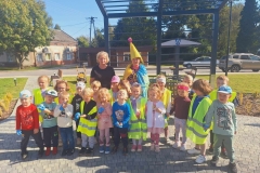 Grupa dzieci stoi w półkolu na szarej kostce. Kilka z nich ma założone kamizelki odblaskowe. Za ich plecami stoi pani w czarnej bluzce i eko- skrzat w żółtej czapce i pelerynie z kolorowych worków na śmieci. Za nimi zielona trawa, obok ławeczka.