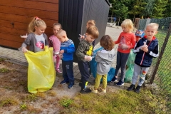 Grupa dzieci stoi przed bramą, dwie dziewczynki trzymają żółty i zielony worek na śmieci. Każde dziecko ma na ręce rękawczkę.