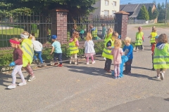 Dzieci zbierają śmieci obok ogrodzenia. Na prawej ręce mają niebieską rękawiczkę. Jeden chłopiec w odblaskowej kamizelce, drugi w niebieskie bluzie i dziewczynka w różowej bluzeczce trzymają niebieski worek. W tle biały dom.