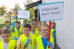 Grupa przedszkolaków stoi w gromadce. Mają na sobie kamizelki odblaskowe. Dwóch chłopców trzyma plastikowe laski z wypisanymi na kartonie hasłami: ,, Dbamy  o planetę’’ oraz ,, Sprzątamy nasz świat’’. Napisy wykonane są czarnymi, drukowanymi literami na białym tle w niebieskim obramowaniu. Jeden z chłopców podnosi zielony worek zapełniony do połowy szkłem. W tle widoczne są drzewa.