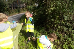 Czworo przedszkolaków ubranych w żółto - srebrne kamizelki odblaskowe porządkuje pas zieleni obok polnej drogi. Jeden z chłopców trzyma żółty worek, dwóch wrzuca do niego śmieci, m. inn. plastikową butelkę po Coca - coli. Czwarty chłopiec przykucnął i podnosi coś z ziemi. W tle zieleń drzew i krzewów.