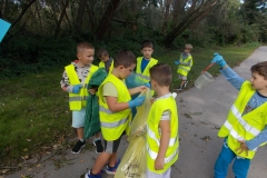 Siedmiu chłopców stoi na polnej drodze. Na swoje kolorowe stroje mają nałożone kamizelki odblaskowe. Zbierają z drogi i przyległego do niej pasa zieleni śmieci segregując je do żółtego i zielonego worka. Jeden z chłopców pokazuje, że znalazł plastikowe wiadereczko. W tle drzewa i zielone krzewy.