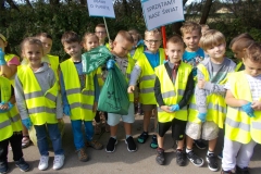 Obok budynku przedszkola stoi grupa dzieci w kamizelkach odblaskowych. Na dłoniach mają niebieskie gumowe rękawice. Dwóch chłopców trzyma plastikowe laski z wypisanymi na kartonie hasłami: ,, Dbamy  o planetę’’ oraz ,, Sprzątamy nasz świat’’. Napisy wykonane są czarnymi, drukowanymi literami na białym tle w niebieskim obramowaniu. W tle widać plac zabaw.