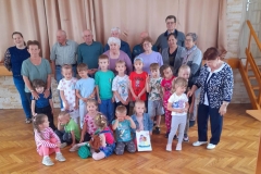 Grupa dzieci stoi wraz z seniorami, kilkoro dzieci siedzi na podłodze.