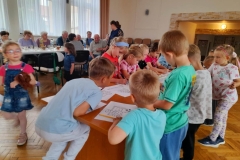 Dzieci i Seniorzy siedzą przy stolikach, dzieci kolorują kolorowanki.