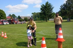 Dwóch strażaków pomaga dziewczynce trzymać węża strażackiego.