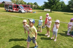 Zdjcie przedstawia grupę dzieci w kolorowych strojach. W tle dwa wozy strażcie.