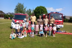 Kilkoro dzieci kuca na trawie, pozostałe stoją za nimi wraz z piecioma dorosłymi. W tle dwa wozy strażackie.