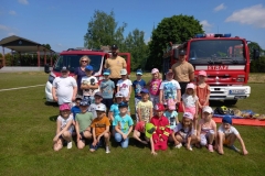 Kilkoro dzieci kuca na trawie, pozostałe stoją za nimi wraz z pięcioma dorosłymi. W tle dwa wozy strażackie.