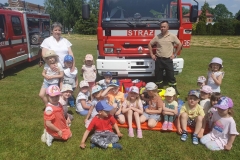 Grupa dzieci siedzi na trawie. Za nimi stoją dwie osoby dorosłe. W tle samochód strażacki.