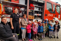 Grupa dzieci stoi przed otwarty wozem strażackim z druhną Julią i druhem Marcinem. W wozie strażackim widać jego wyposażenie.