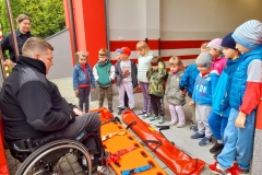 Druh Marcin tłumaczy dziecim jak umieszcza się chorego na noszach, które są rozłożone na ziemi.