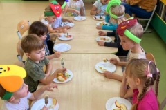 Dzieci siedzą przy stolikach, każde z nich na talerzyku ma owoce I warzywa, spożywają je. W tle widać rodziców.