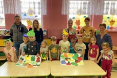 Dzieci wraz z rodzicami stoją za stolikami, trzymają w rękach wykonane plansze z owocami i warzywami.