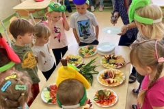 Grupa dzieci zgromadzona wokół stołu, na stole kilka talerzy z pokrojonymi owocami I warzywami.