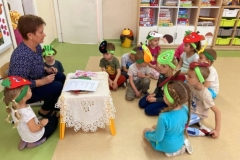 Grupa dzieci siedzi na podłodze, na głowach mają kolorowe opaski z owocami i warzywami, przed nimi stolik przy którym siedzi pani. Po obu stronach pani siedzą przedszkolaki.