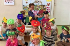 Uśmiechnięte dzieci siedzą na podłodze, na głowie mają kolorowe opaski z owocami i warzywami. Za nimi pani, trzyma w ręce postać wróżki. W tle tablica z piramidą zdrowia.